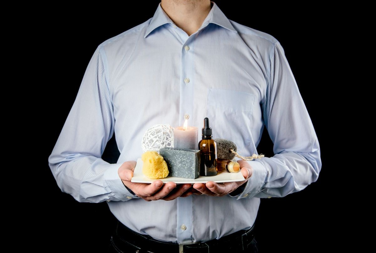Manly businessman holding white ceramic tray with group of different spa essentials, beard oil, gray soap, natural sea sponge, candle burning, aroma oil on black background. Man spa concept. 