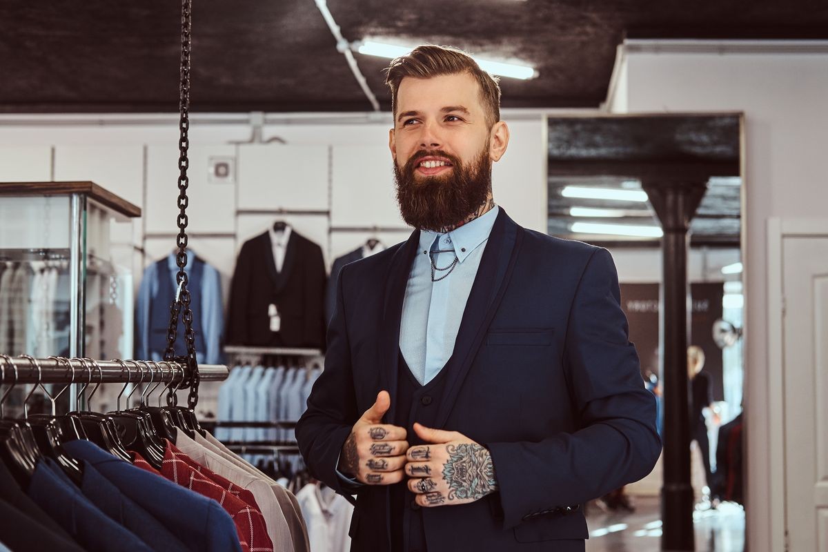 Smiling tattoed male with stylish beard and hair dressed in elegant suit standing in menswear store.